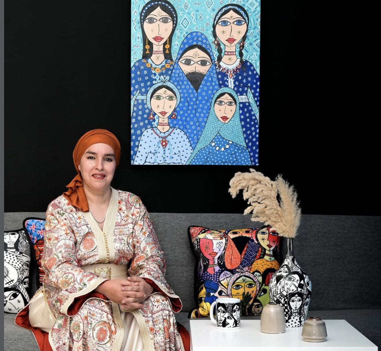 Hafida Zizi seated beneath one of her paintings, surrounded by objects carrying her work